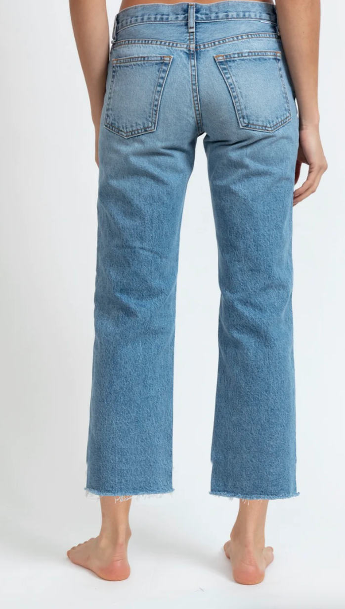 A person from behind wearing ASKK&#39;s berkeley low rise straight cropped blue jeans with frayed hems in Arizona style, standing barefoot on a white background.
