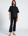 A woman stands against a plain background in a Scottsdale Arizona bungalow, wearing a loose-fitting black ASKK boy tee and dark cropped jeans, barefoot, looking directly at the camera with a subtle expression.