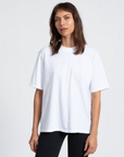 A woman with shoulder-length brown hair stands wearing a plain white ASKK boy tee and black pants against a light gray background in Scottsdale, Arizona. She looks directly at the camera with a neutral expression.