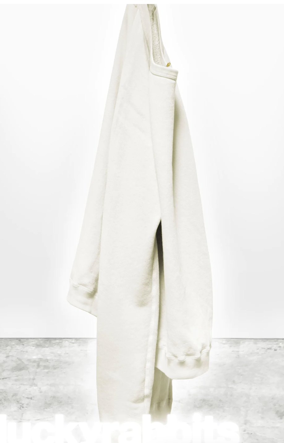 A pair of cream-colored, oversized Lucky Rabbits sweatpants hanging from a hook in a bungalow in Scottsdale, Arizona against a white wall with a gray concrete floor, displaying the text "lucky rabbits" near the bottom. Brand: Free City (sparrow, LLC)