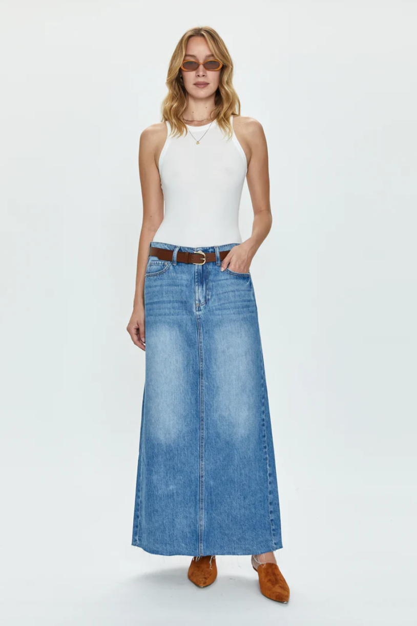A woman in a white tank top and the RAYE MAXI SKIRT - HIGH NOON denim skirt with a brown belt, standing on a plain background, wearing brown ankle boots and sunglasses from the Los Angeles-based womenswear brand, Pistola.