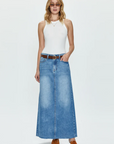 A woman in a white tank top and the RAYE MAXI SKIRT - HIGH NOON denim skirt with a brown belt, standing on a plain background, wearing brown ankle boots and sunglasses from the Los Angeles-based womenswear brand, Pistola.
