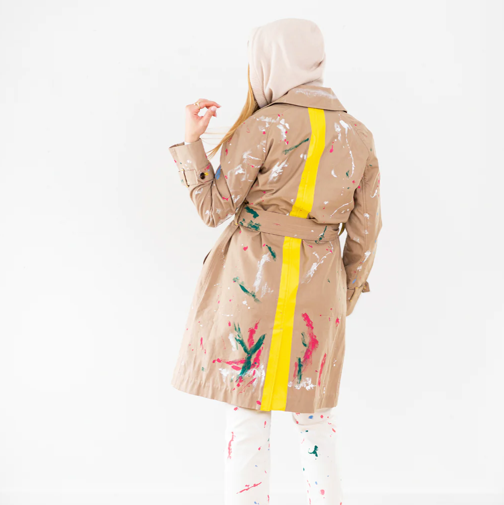 A woman in a hijab, viewed from behind, wears a paint-splattered beige Kerri Rosenthal trench coat with a yellow belt and colorful splattered pants against a white background in Scottsdale, Arizona.