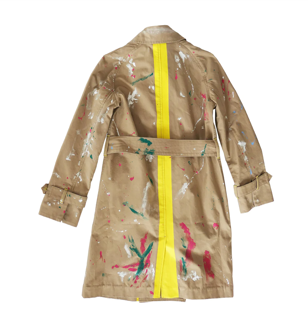 A beige trench coat with random colorful paint splatters and a bright yellow vertical stripe at the back, displayed against a white background in a Scottsdale bungalow by Kerri Rosenthal's The KR Trench.