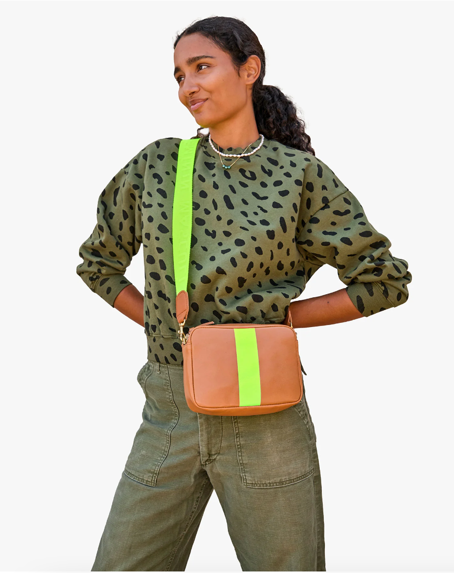 A woman wearing olive green jeans and a leopard print top with a Clare Vivier neon strap crossbody bag smiles, posing against a plain background. Her style perfectly complements the laid-back Arizona aesthetic.