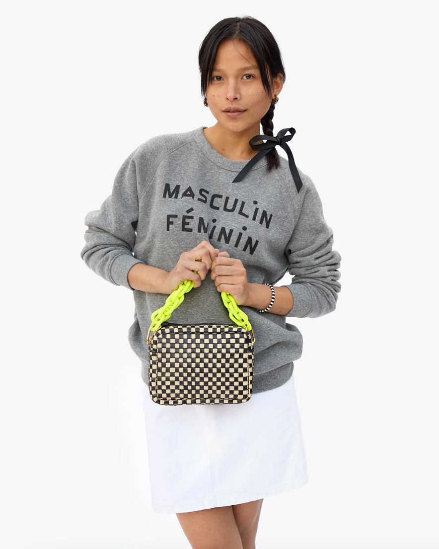 A person with long dark hair tied with a black ribbon is wearing a grey sweatshirt with &quot;MASCULIN FÉMININ&quot; written on it and a white skirt. They are holding a checkered handbag with neon yellow handles and an attached Clare Vivier Shortie Strap in front of them. The background is plain white.