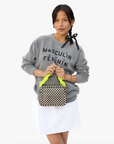 A person with long dark hair tied with a black ribbon is wearing a grey sweatshirt with "MASCULIN FÉMININ" written on it and a white skirt. They are holding a checkered handbag with neon yellow handles and an attached Clare Vivier Shortie Strap in front of them. The background is plain white.