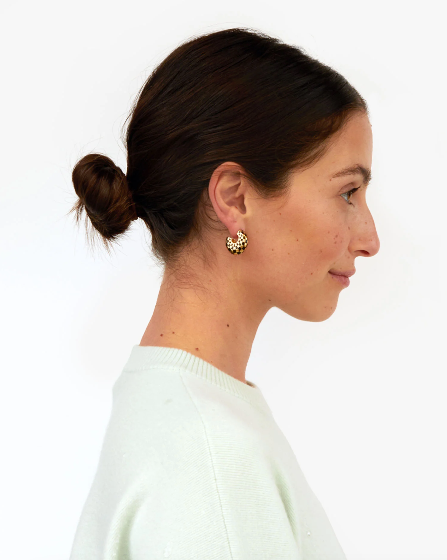 Profile view of a woman with her hair in a neat bun, wearing a white sweater and gold earrings, showcasing Le Hoop Black &amp; Gold Checker style by Clare Vivier against a white background.