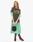 A person with long red hair stands against a white background, wearing a gray T-shirt featuring a yellow fish design over a green and white striped dress, paired with brown boots. They hold the "Pot de Miel" handbag by Clare Vivier, featuring bamboo handles.