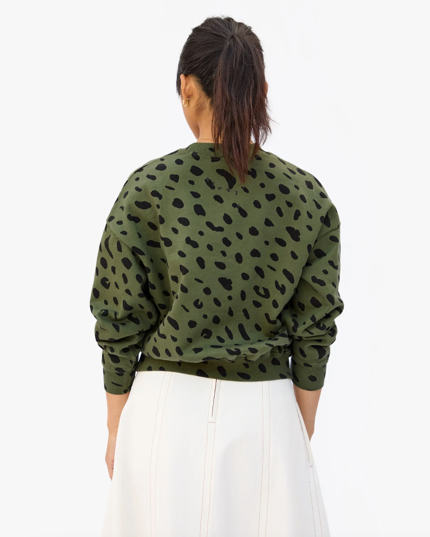 A woman viewed from behind, wearing a green Le Drop Army sweater by Clare Vivier with black jaguar print and white trousers, embodies Arizona style. Her hair is tied back in a ponytail.