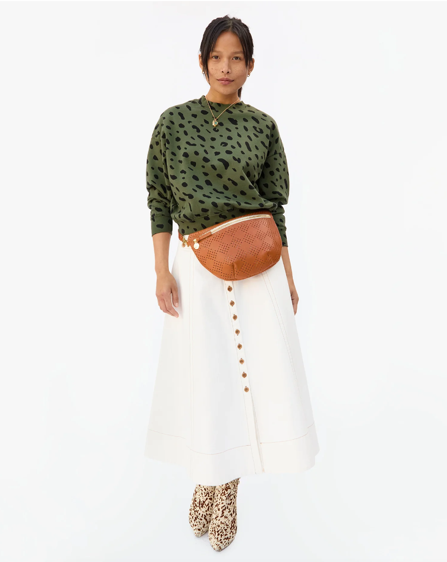 A person standing against a white background, wearing a green leopard print top, white skirt with button details, Le Drop Army w/ Black Jaguar boots from Clare Vivier, and a tan crossbody bag.