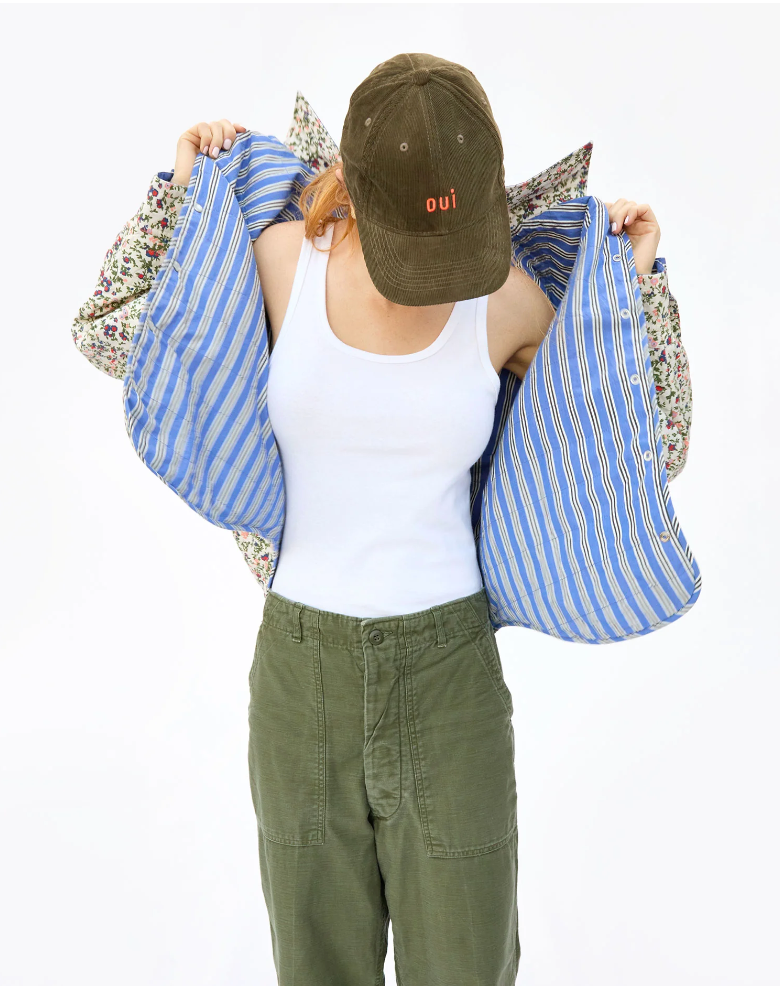 A woman wearing a Baseball Hat Olive Corduroy Oui by Clare Vivier faces away from the camera, holding up two large, colorful tote bags by their handles. Her attire includes a white tank top and Arizona green trousers.