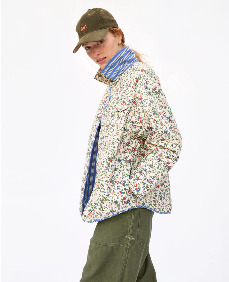A woman wearing a floral jacket, blue striped scarf, Clare Vivier baseball hat in Olive Corduroy with "DIY" label, and olive pants looks over her shoulder against a white background, exuding a unique Arizona style.