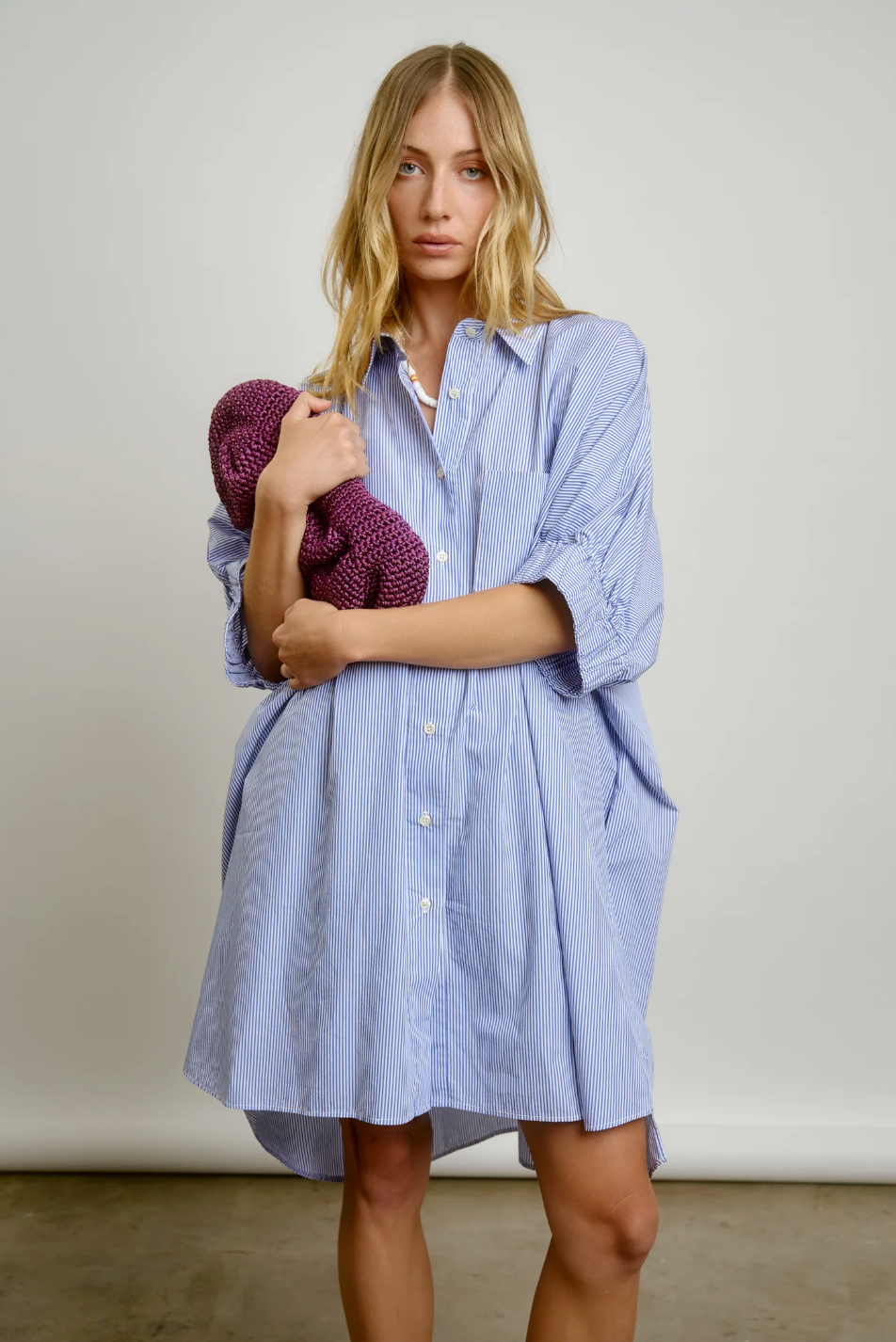 A woman with blonde hair stands holding a purple knitted hat, dressed in a light blue striped Le Shirt dress in front of a neutral background in her Scottsdale, Arizona bungalow by Aquarius Cocktail.
