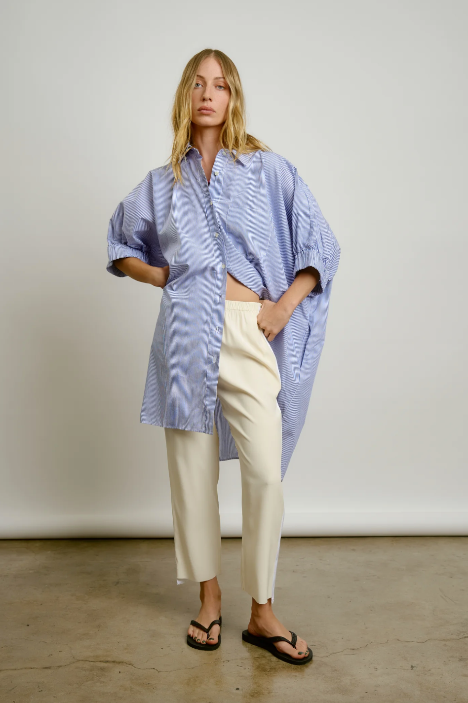 A woman stands in a studio in Scottsdale, Arizona, wearing a blue oversized striped Le Shirt from Aquarius Cocktail and cream trousers, paired with black sandals. She has a relaxed pose with one hand on her hip and a neutral expression.
