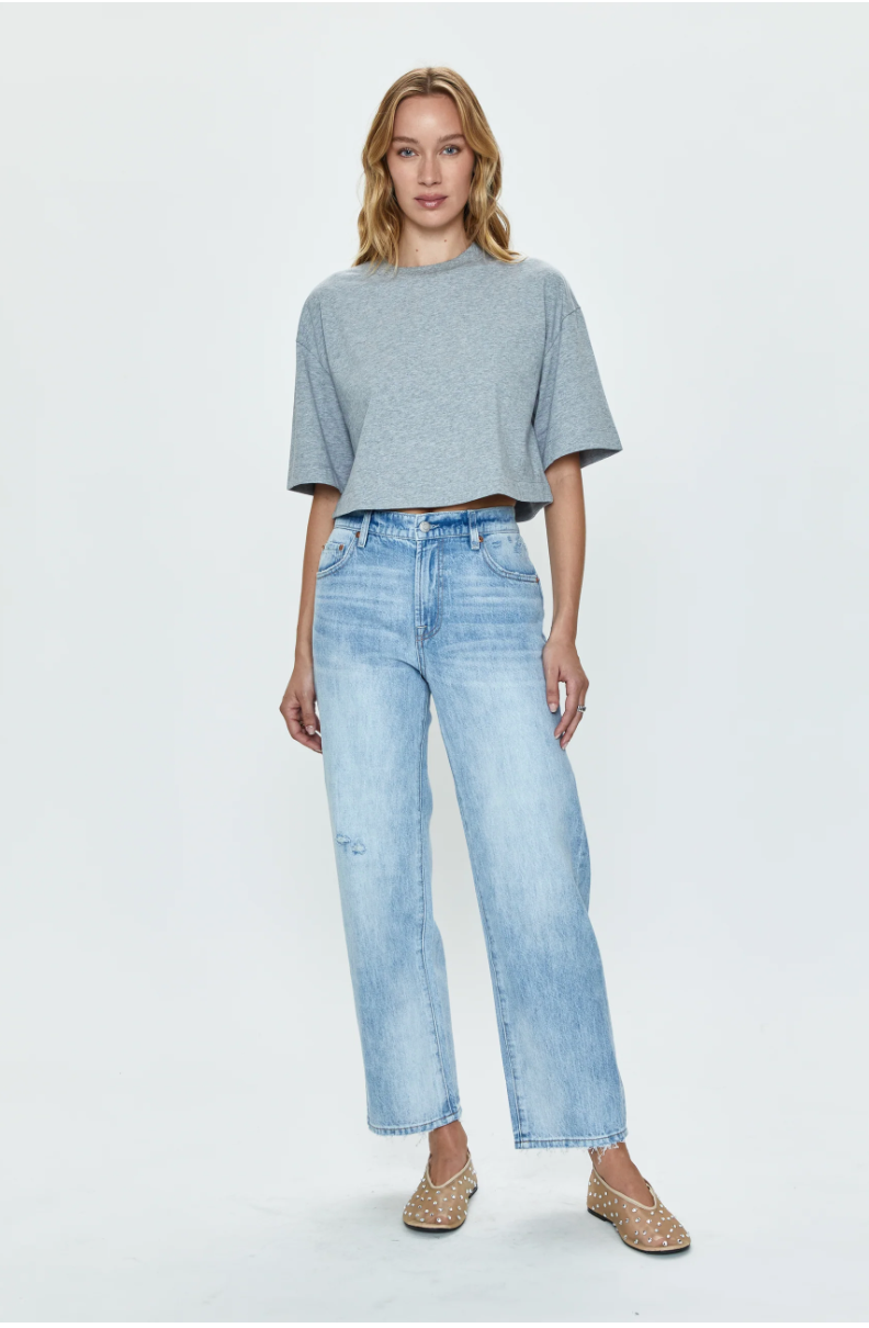 A woman standing in a studio, wearing a Pistola Mae Cropped Tee and light blue ripped jeans, paired with embellished flat shoes, against a plain white background.