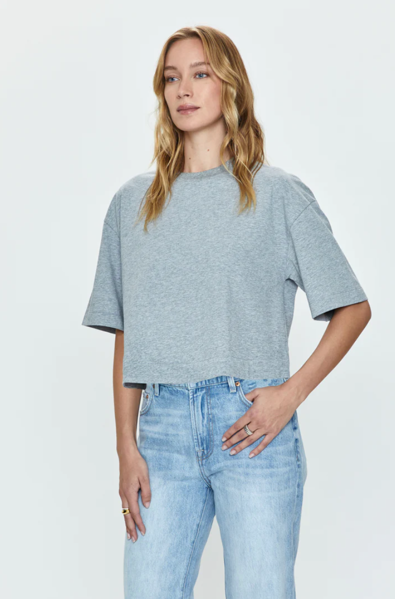 A Los Angeles-based woman in a Pistola Mae Cropped Tee and light blue jeans stands against a white background, her hand resting on her hip.