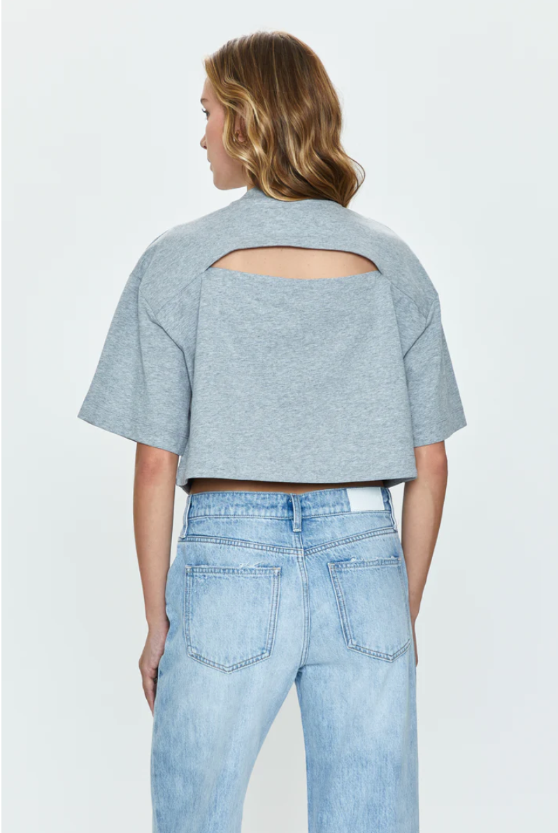 A woman seen from the back wearing a Pistola Mae Cropped Tee with an oval cutout at the upper back, paired with light blue jeans. Her hair is styled in loose waves, capturing a Los Angeles-based style.