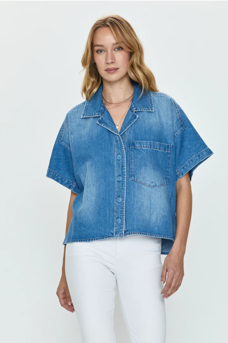 A woman wearing a loose-fitting blue denim Della Camp shirt by Pistola with short cuffed sleeves and a large front pocket, paired with white pants, showcases her Los Angeles-based style against a plain background.