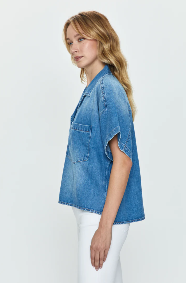 A woman in a blue denim Della Camp shirt from Pistola and white pants, viewed from the side, glances over her shoulder with her blonde hair falling softly around her face, embodying effortless Los Angeles-based style.