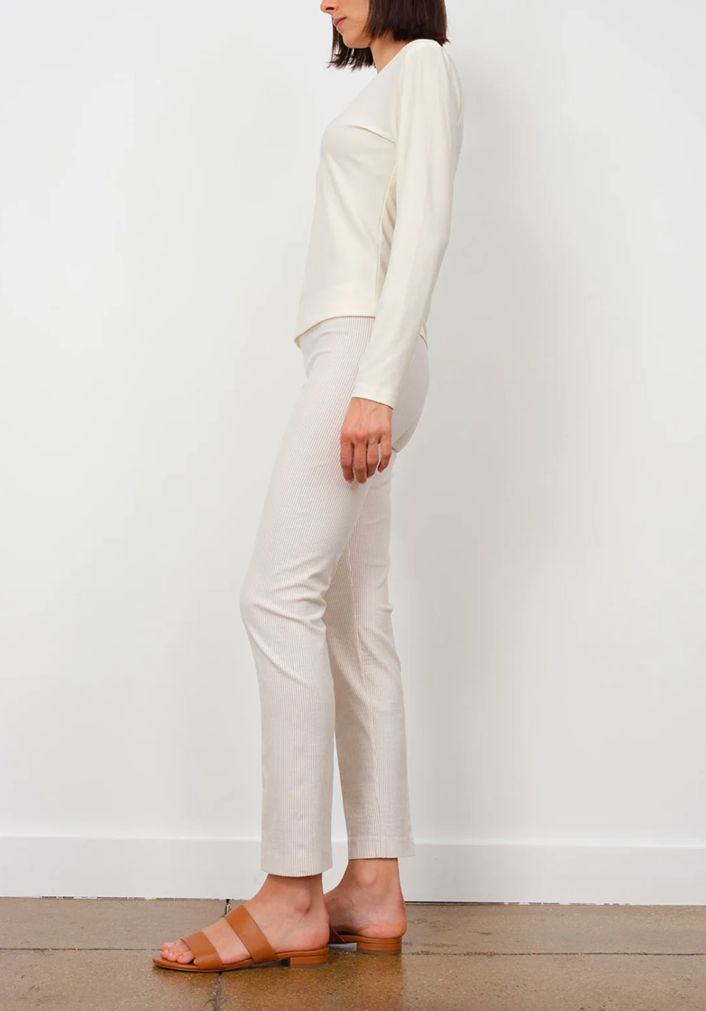 A woman stands against a plain background wearing Avenue Montaigne&#39;s Lulu Pant in cream, paired with a matching long-sleeve top and brown open-toe sandals. The image, captured in Scottsdale Arizona, shows her from the side.