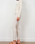 A woman stands against a plain background wearing Avenue Montaigne's Lulu Pant in cream, paired with a matching long-sleeve top and brown open-toe sandals. The image, captured in Scottsdale Arizona, shows her from the side.