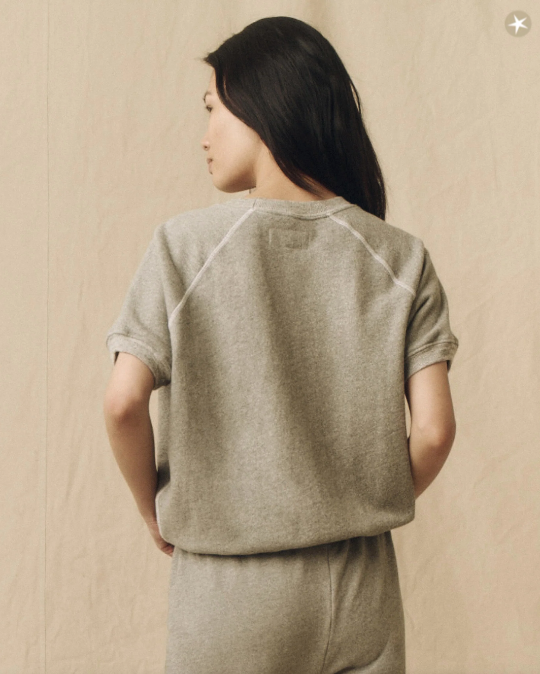 A woman with long black hair wearing The Short Sleeve Sweatshirt Varsity Grey from The Great Inc., seen from behind, against a beige background in Scottsdale Arizona.