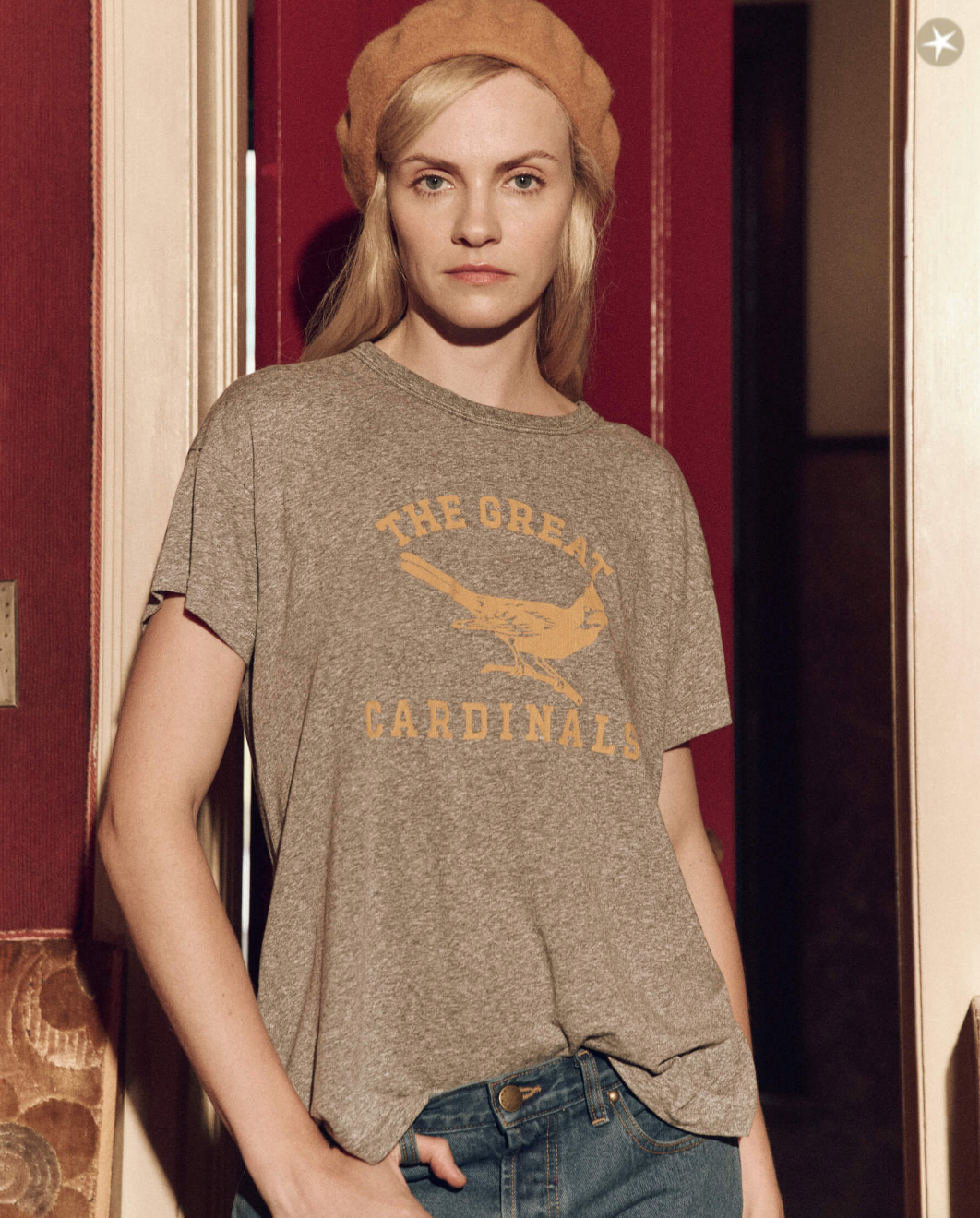 A woman in a grey The Boxy Crew w/ Perched Cardinal t-shirt from The Great Inc. with a bird illustration, wearing a beige beret, stands in a warm-toned, elegant bungalow.