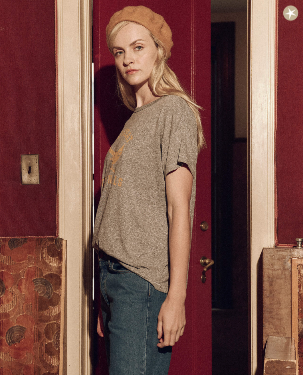A young woman in a casual grey The Boxy Crew w/ Perched Cardinal t-shirt and blue jeans, wearing a mustard beret, stands in the doorway of a Scottsdale bungalow with maroon walls. She poses with a gentle gaze directed off-camera.