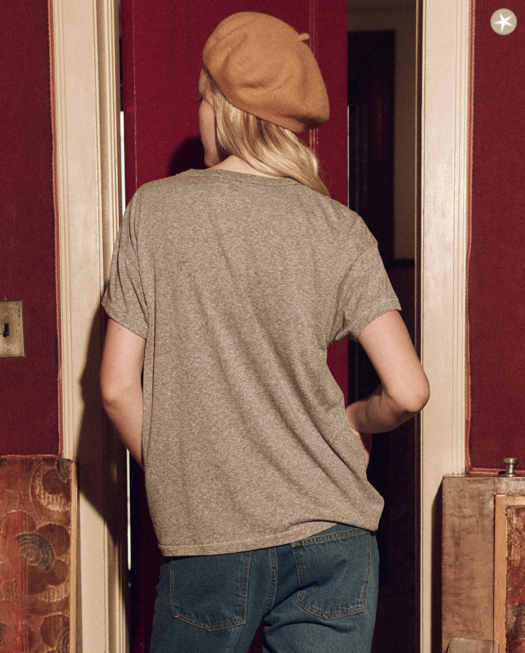 A person standing with their back to the camera, wearing The Boxy Crew w/ Perched Cardinal t-shirt by The Great Inc., blue jeans, and a tan beret, is seen entering a bungalow with red walls and a vintage feel.