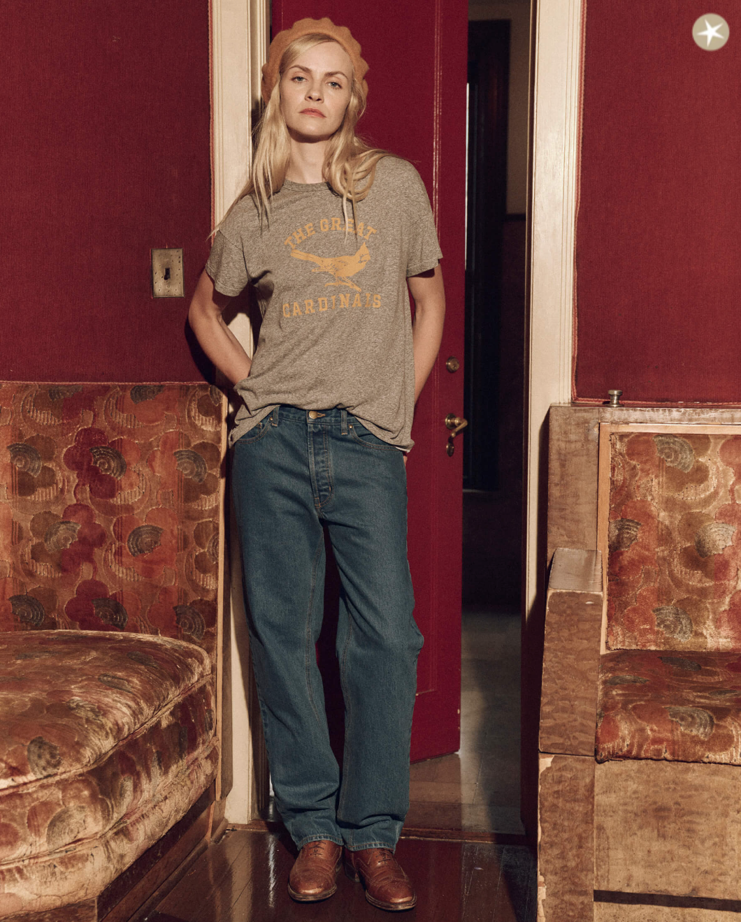 A woman with blonde hair stands confidently in a bungalow, wearing a gray vintage-style The Boxy Crew w/ Perched Cardinal T-shirt by The Great Inc., blue jeans, and brown leather shoes, with her hands in her pockets. She is surrounded by old-fashioned red upholstered chairs.