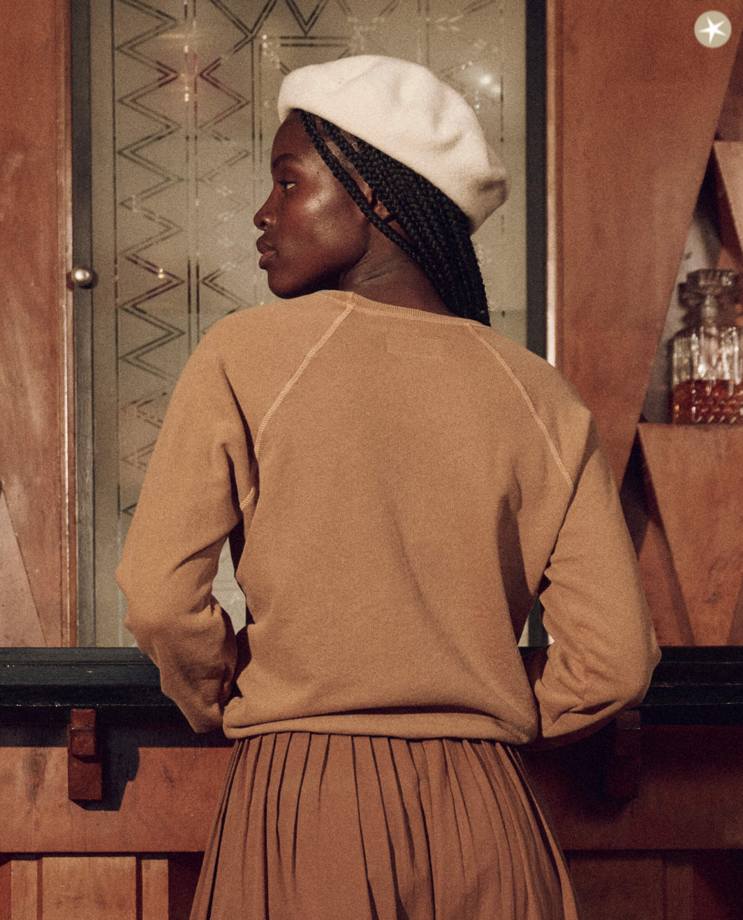 A woman in a cozy The Shrunken Sweatshirt and skirt, with a white beret, looks pensively to the side. She stands indoors near a wooden counter in a Scottsdale, Arizona bungalow, and a wall with decorative motifs.
