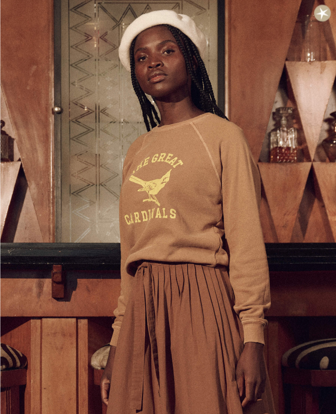 A Black woman wearing a white beret and The Shrunken Sweatshirt from The Great Inc. with the WASHED SUNTAN WITH PERCHED CARDINAL GRAPHIC print stands in front of a wooden and glass background of a Scottsdale, Arizona bungalow, looking thoughtfully to her left.