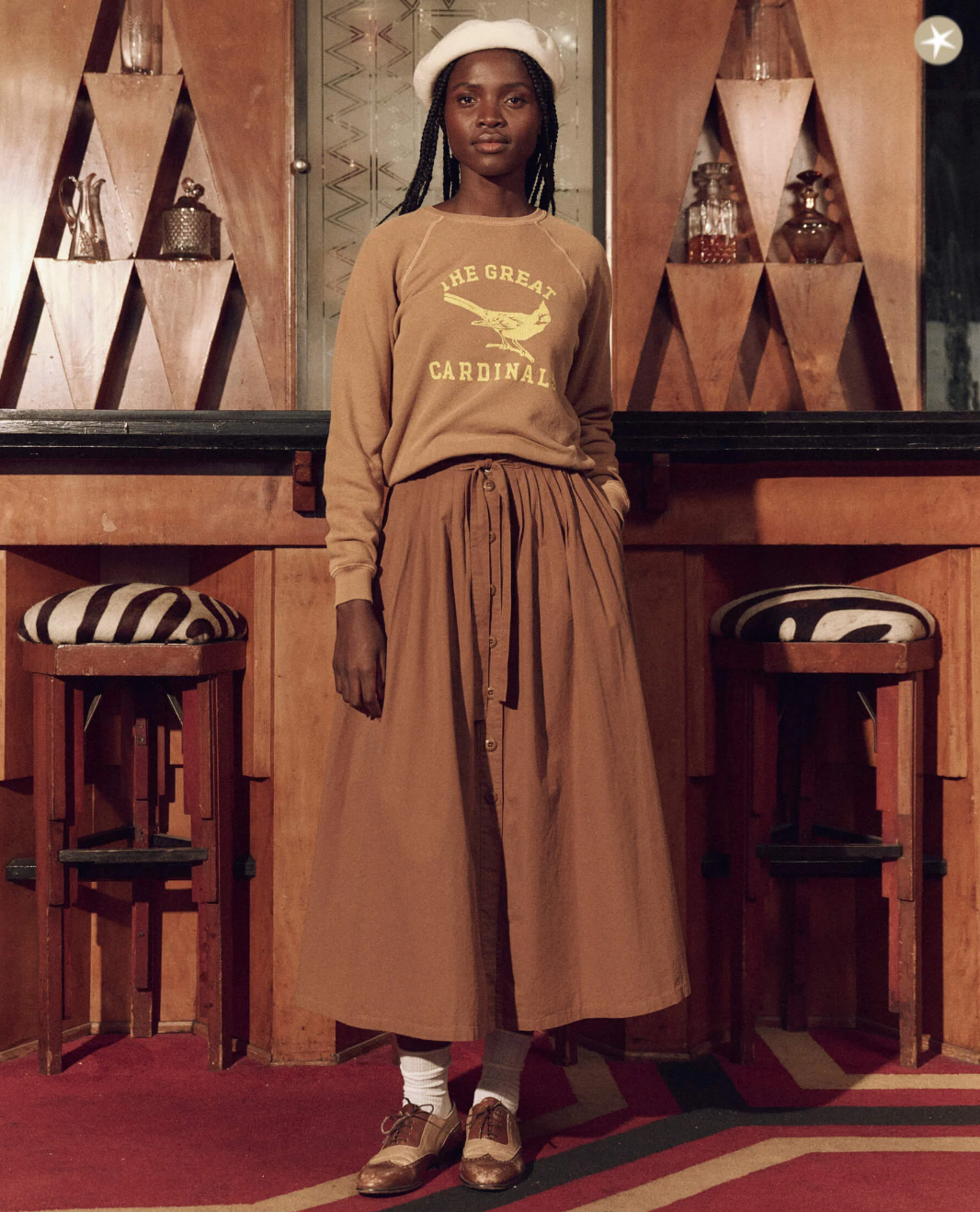 A woman stands in a classic, bungalow-style room wearing a brown midi skirt, The Shrunken Sweatshirt in Washed Suntan with Perched Cardinal graphic by The Great Inc., a cap, and white socks with brown loafers.