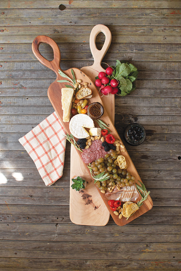 A Pecan Charcuterie Board by Kalalou, Inc filled with assorted cheeses, cold cuts, fruits, and nuts, arranged on a rustic wooden table at a Scottsdale, Arizona bungalow, accompanied by a napkin and a glass of red wine.