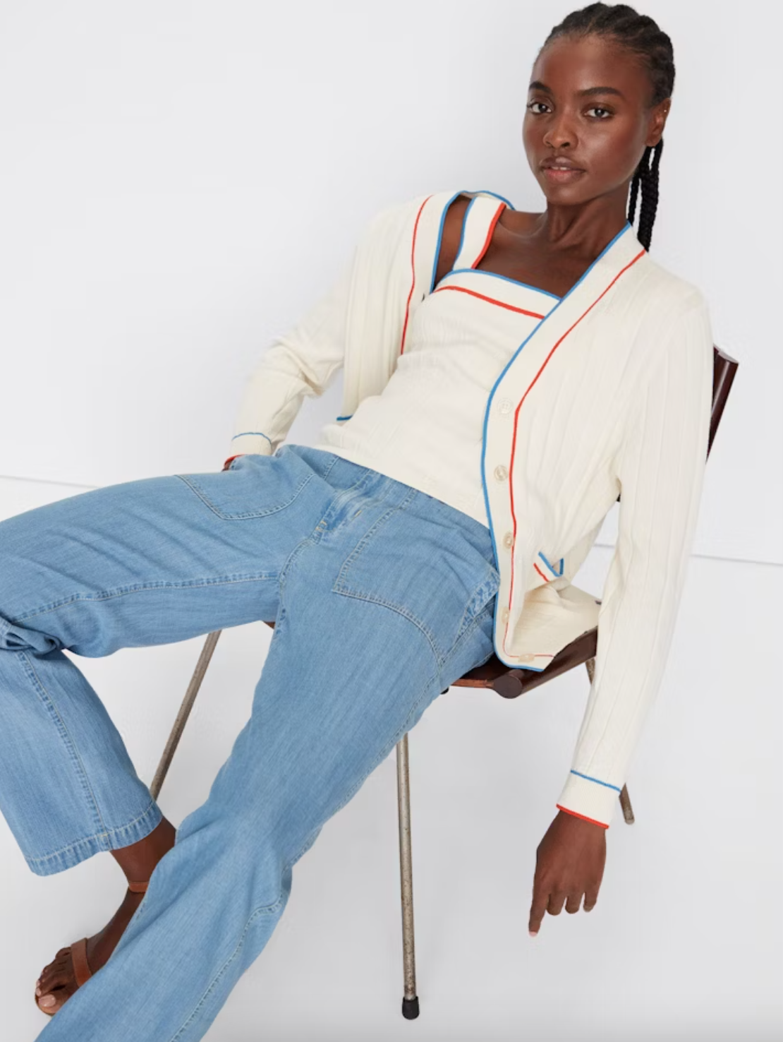 A Black woman in stylish casual attire sits casually on a metal chair in Scottsdale, Arizona. She wears a white striped cardigan over an orange and blue trimmed top, paired with light blue jeans and brown sandals from Kule's THE DEDE CREAM collection.