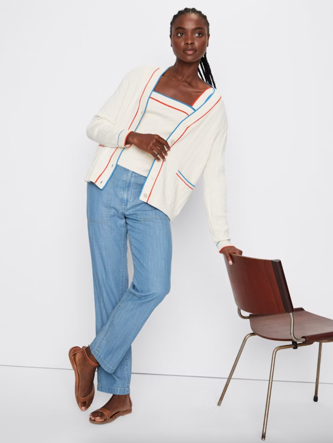 A stylish young woman in a white cardigan and blue jeans, leaning casually against a chair in her Scottsdale, Arizona bungalow. She wears brown open-toe heels and her hair in braids while applying THE DEDE CREAM by Kule.