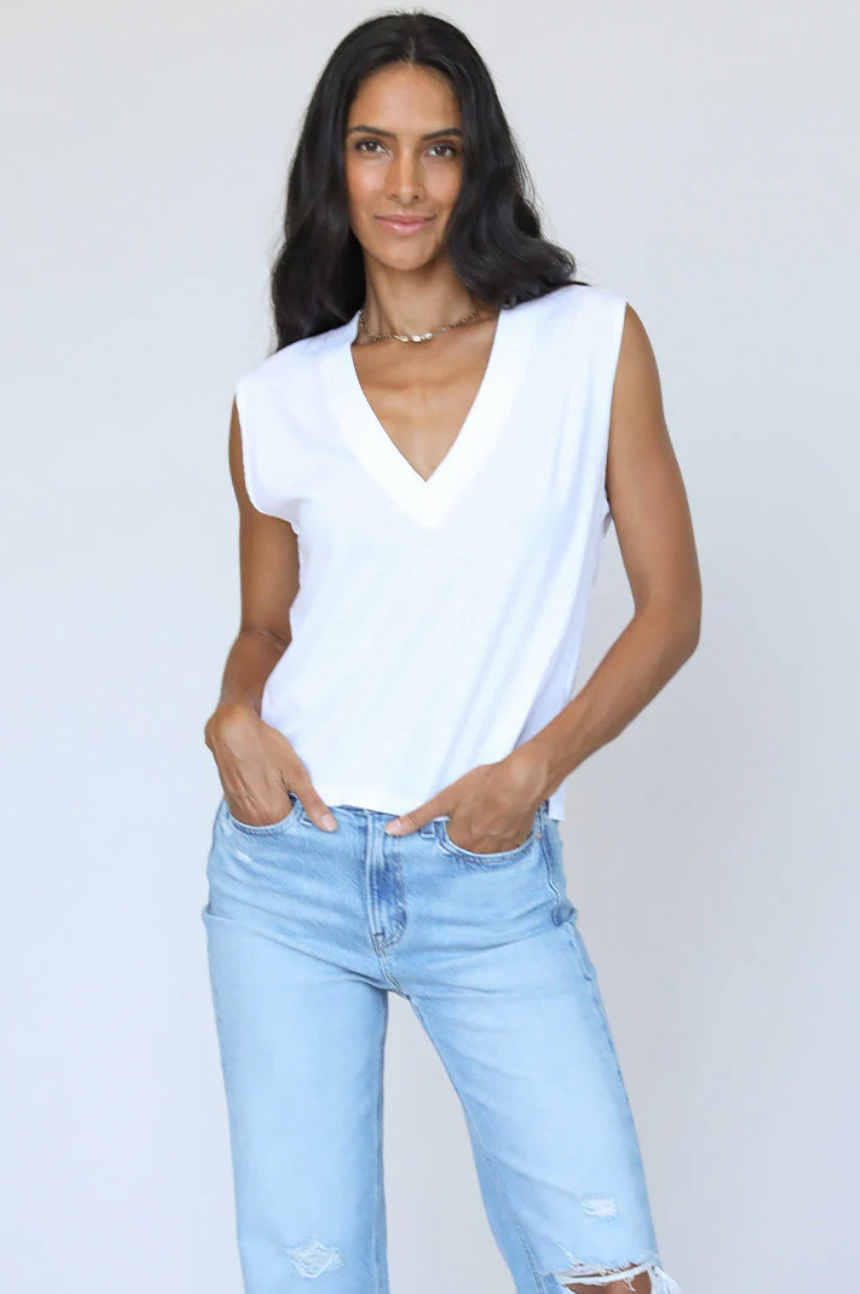 A woman wearing a white Margot Cotton Sleeveless V-Neck Tee and distressed blue jeans stands against the plain background of a bungalow, hands lightly resting on her hips, smiling slightly.