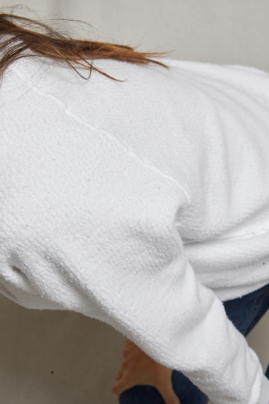 Close-up view of a person wearing a Perfectwhitetee Ziggy Reverse Fleece LS Shrunken Crewneck Sweatshirt and dark jeans, seated outside a bungalow with their back turned partially to the camera. The focus is on the texture of the sweatshirt.