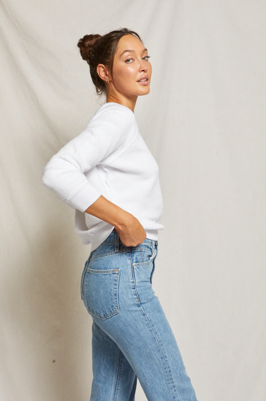 A woman in a white Ziggy Reverse Fleece LS Shrunken Crewneck Sweatshirt and blue jeans looks over her shoulder with a slight smile against the neutral background of a bungalow. She has her hair in a bun and adopts a casual pose.