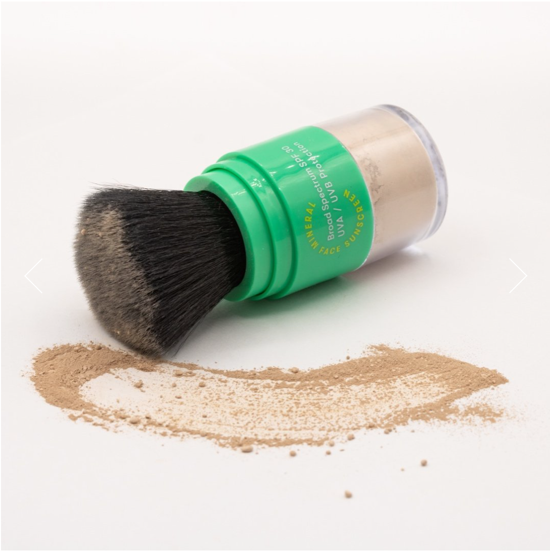 A makeup brush with a green handle, lying on its side, with loose Larkly SPF 30 Mineral Powder Sunscreen spilled around it on a white background in Scottsdale, Arizona.