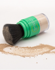 A makeup brush with a green handle, lying on its side, with loose Larkly SPF 30 Mineral Powder Sunscreen spilled around it on a white background in Scottsdale, Arizona.