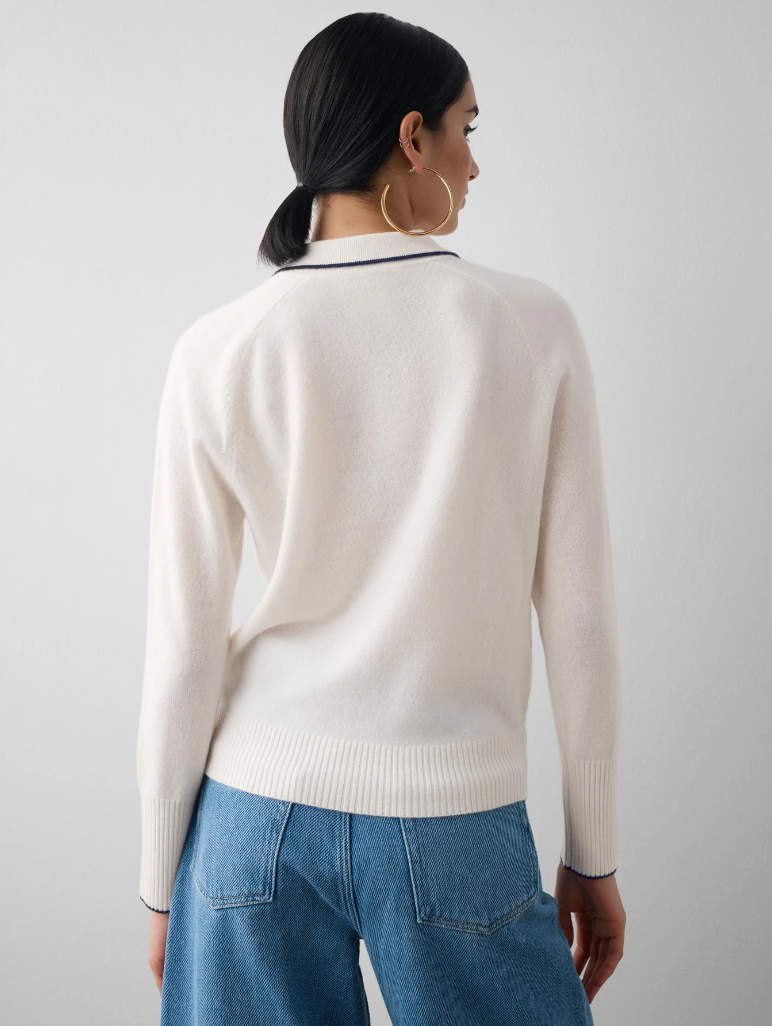 A woman viewed from the back, wearing a chic White + Warren cashmere ribbed trim polo and blue jeans, accessorized with large hoop earrings, stands in front of a charming bungalow in Scottsdale, Arizona. The sweater has a dark stripe along the neckline.