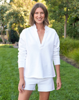 A woman with shoulder-length hair smiling, wearing Frank & Eileen's PEARL Favorite Sweatshorts TRIPLE FLEECE White and a white long-sleeved blouse, standing in the garden of a Scottsdale Arizona bungalow with lush greenery in the background.