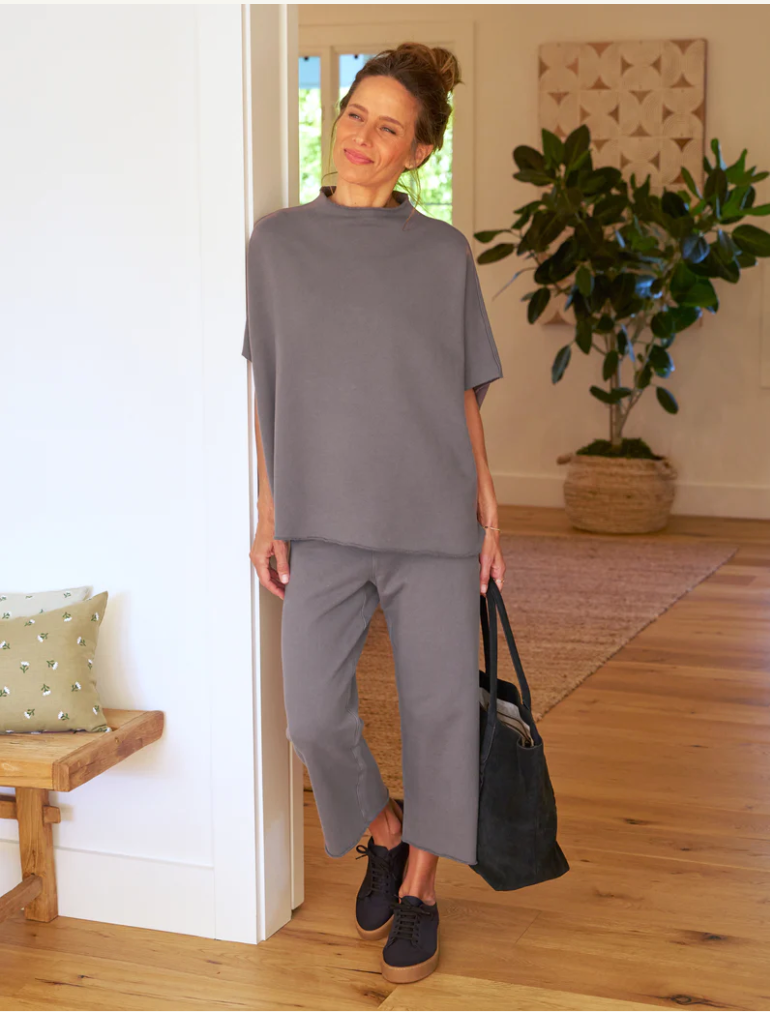 A woman in a casual grey outfit smiles and poses in the warmly lit hallway of a Scottsdale, Arizona bungalow, holding a dark handbag, with a neutral-tone decor background while wearing the CATHERINE Favorite Sweatpant TRIPLE FLEECE by Frank &amp; Eileen.