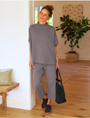 A woman in a casual grey outfit smiles and poses in the warmly lit hallway of a Scottsdale, Arizona bungalow, holding a dark handbag, with a neutral-tone decor background while wearing the CATHERINE Favorite Sweatpant TRIPLE FLEECE by Frank & Eileen.