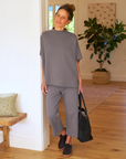 A woman in a casual grey outfit smiles and poses in the warmly lit hallway of a Scottsdale, Arizona bungalow, holding a dark handbag, with a neutral-tone decor background while wearing the CATHERINE Favorite Sweatpant TRIPLE FLEECE by Frank & Eileen.