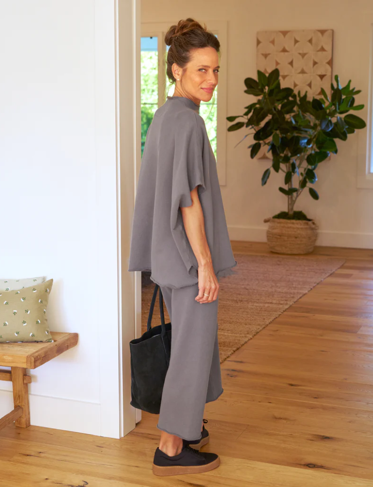 A woman in a stylish gray outfit with Frank & Eileen's CATHERINE Favorite Sweatpant TRIPLE FLEECE and matching pants smiles over her shoulder as she walks through a Scottsdale Arizona bungalow with wood flooring and a potted plant in the background.