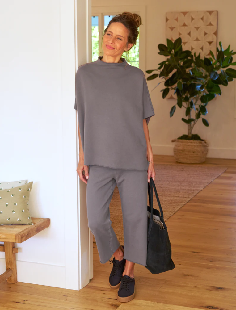 A woman in a casual gray Frank &amp; Eileen AUDREY Funnel Neck Capelet TRIPLE FLEECE outfit stands in a bright room holding a large black bag, smiling gently with indoor plants visible in the background.