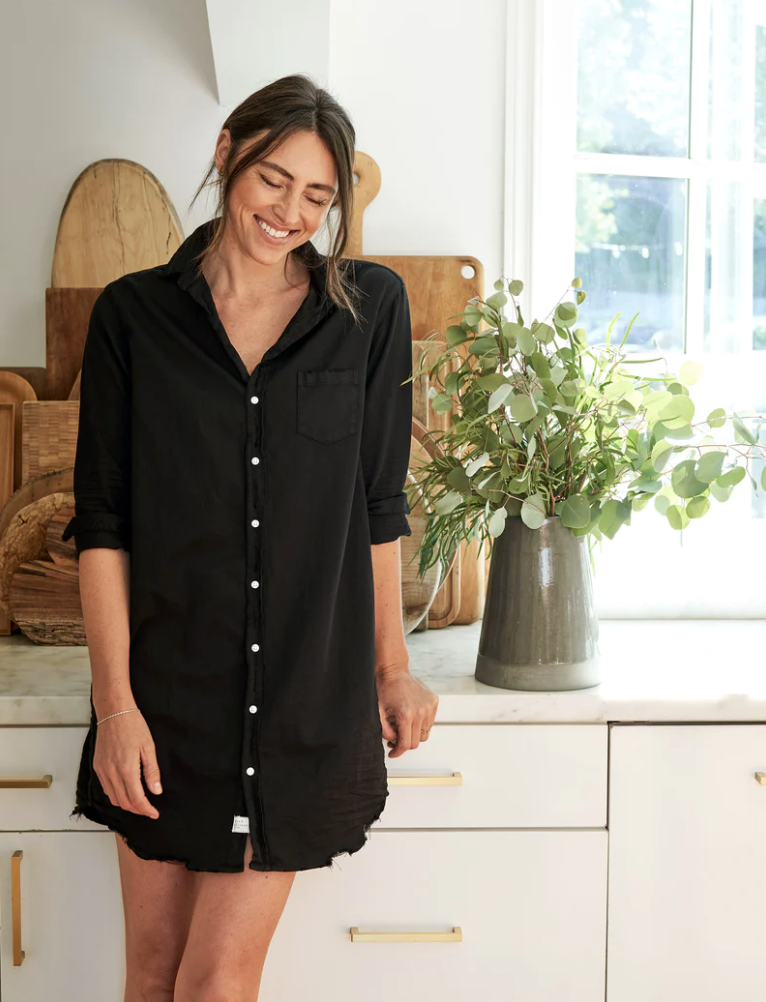 A woman in a black Mary Classic Shirtdress from Frank & Eileen smiles warmly, standing in the bright kitchen of her Scottsdale, Arizona bungalow with wooden accents and a vase of greenery on the countertop beside her.